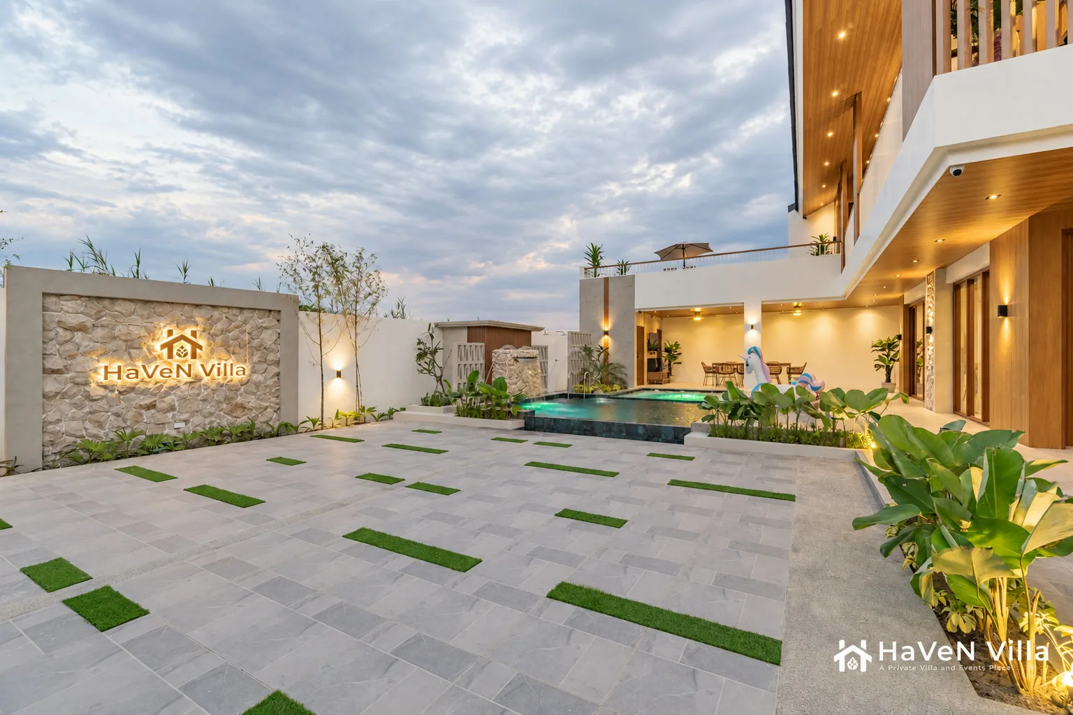 Dusk view across paved courtyard and green inlays toward the pool and warm-lit villa terraces